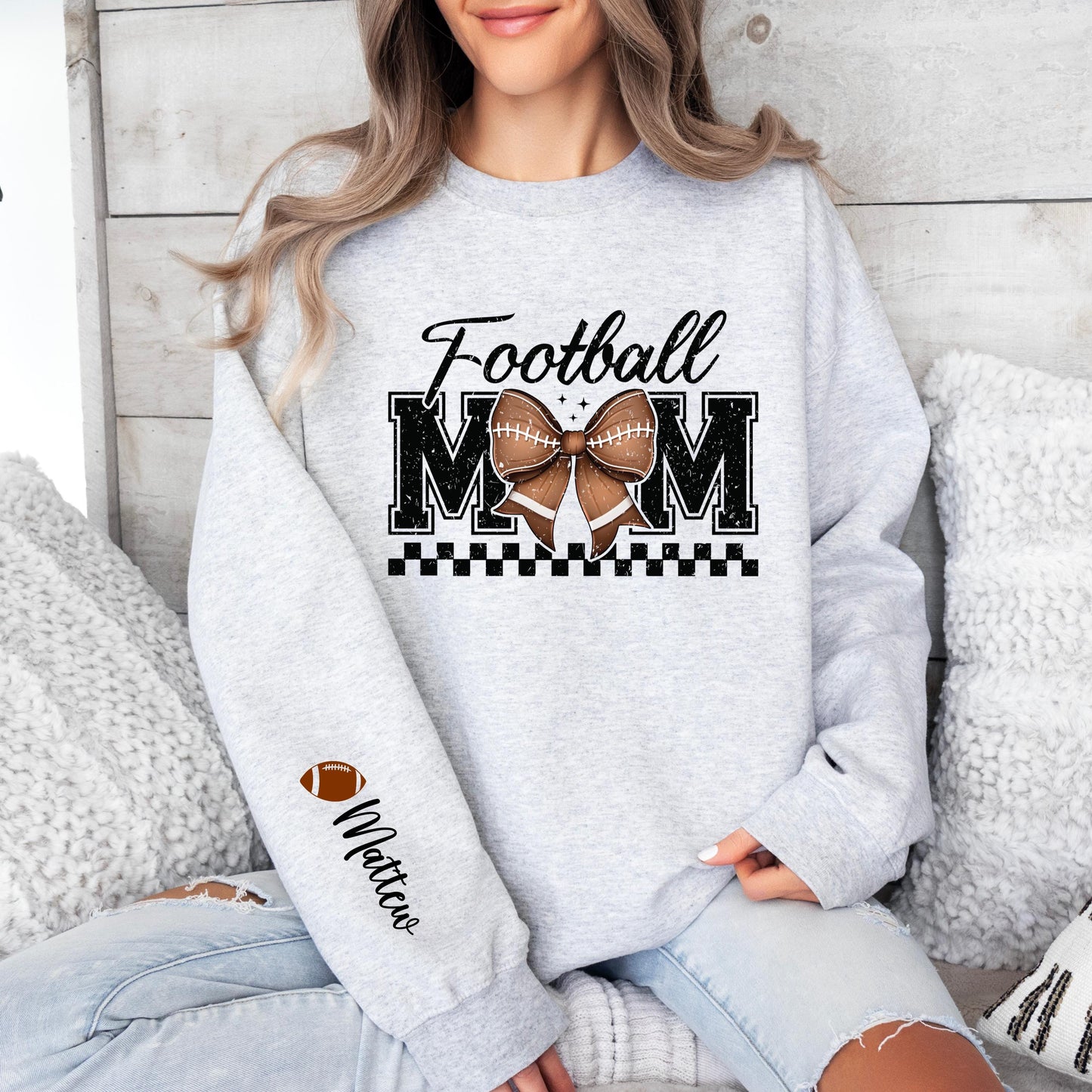 A light gray sweatshirt with "Football MOM" in bold black letters, a football-patterned bow in the center, a black checkered underline, and a football icon with "Custom Name" written on the sleeve.