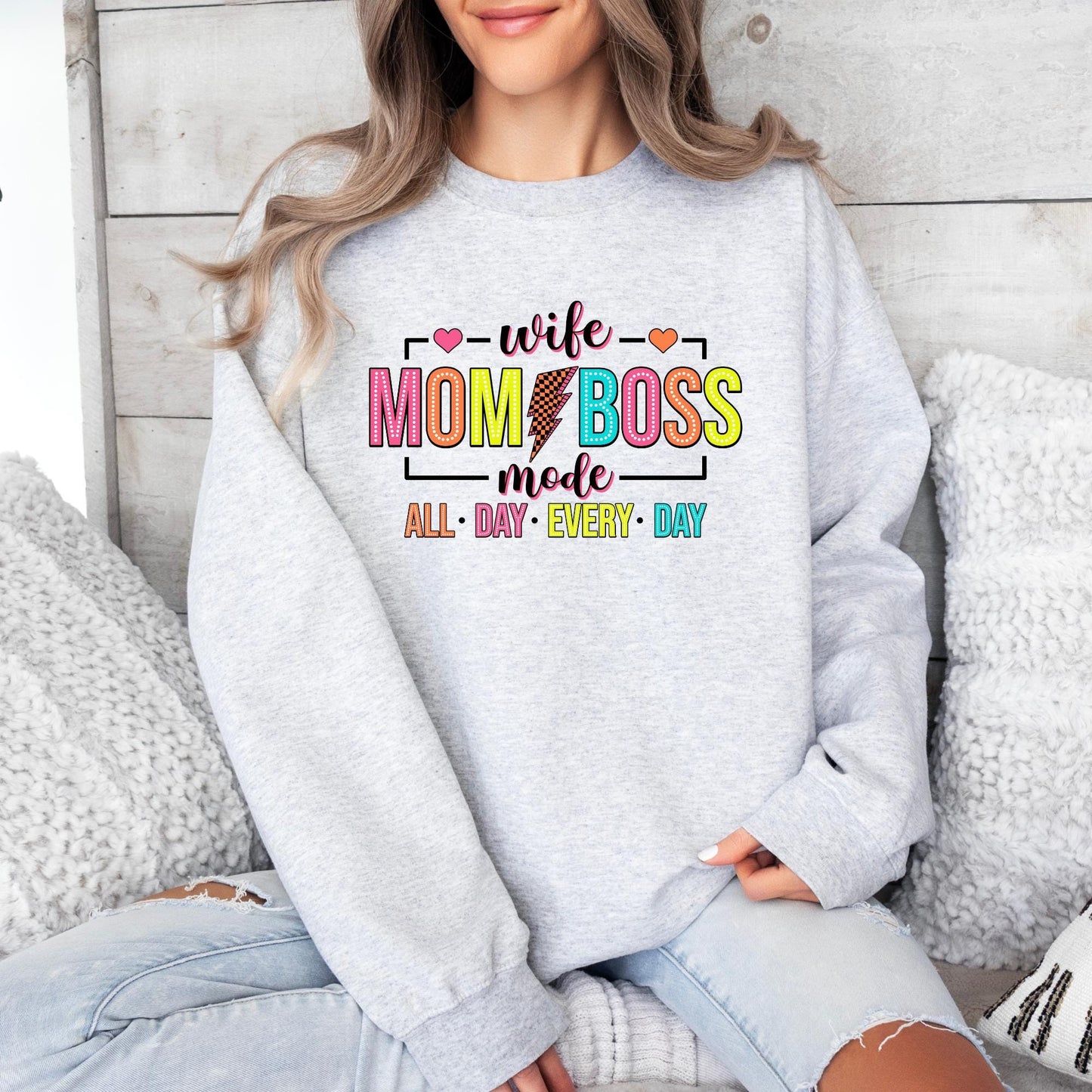 White sweatshirt with "Wife Mom Boss Mode" in bold, colorful letters, featuring a lightning bolt and hearts. The phrase "All Day Every Day" is in bright, playful colors for a fun and empowering look.