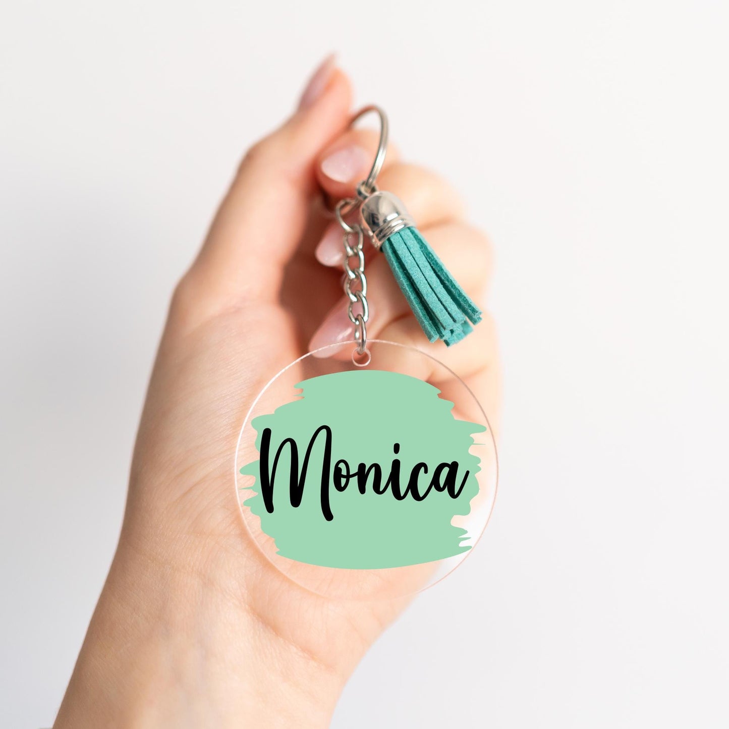 Personalized acrylic keychain with a black name on a pastel green paint stroke design, attached to a silver keyring with a matching green suede tassel.