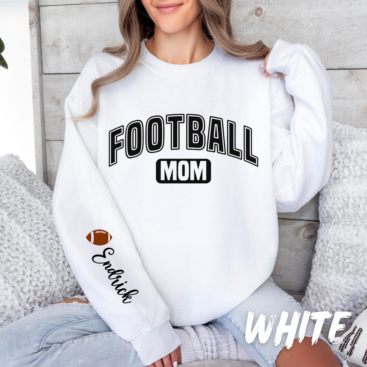 A women sweatshirt with bold black text reading "FOOTBALL MOM" on the front. The left sleeve features a small football graphic and the custom name in cursive.