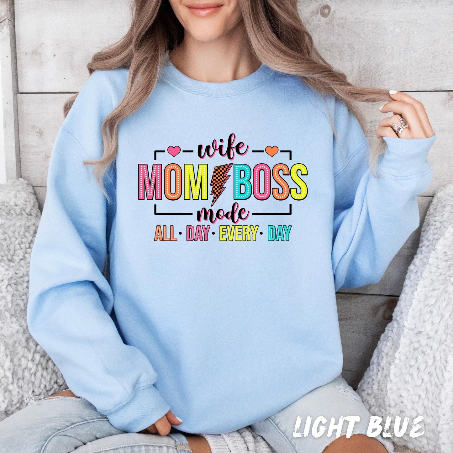 White sweatshirt with "Wife Mom Boss Mode" in bold, colorful letters, featuring a lightning bolt and hearts. The phrase "All Day Every Day" is in bright, playful colors for a fun and empowering look.