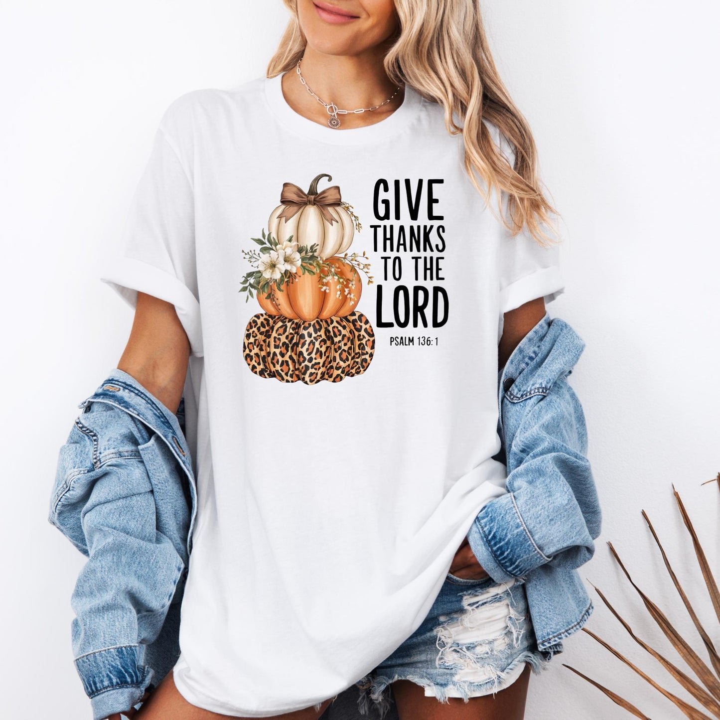 t-shirt with three stacked pumpkins: a leopard-print pumpkin at the bottom, an orange pumpkin in the middle decorated with white flowers, and a cream pumpkin with a brown bow on top. Beside the pumpkins, the text reads “GIVE THANKS TO THE LORD – Psalm 136:1.”