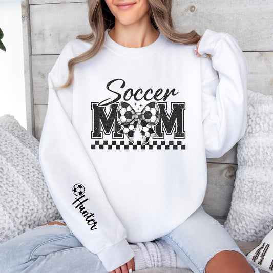 A light gray sweatshirt with &quot;Soccer MOM&quot; in bold black letters, a Soccer -patterned bow in the center, a black checkered underline, and a football icon with &quot;Custom Name&quot; written on the sleeve.