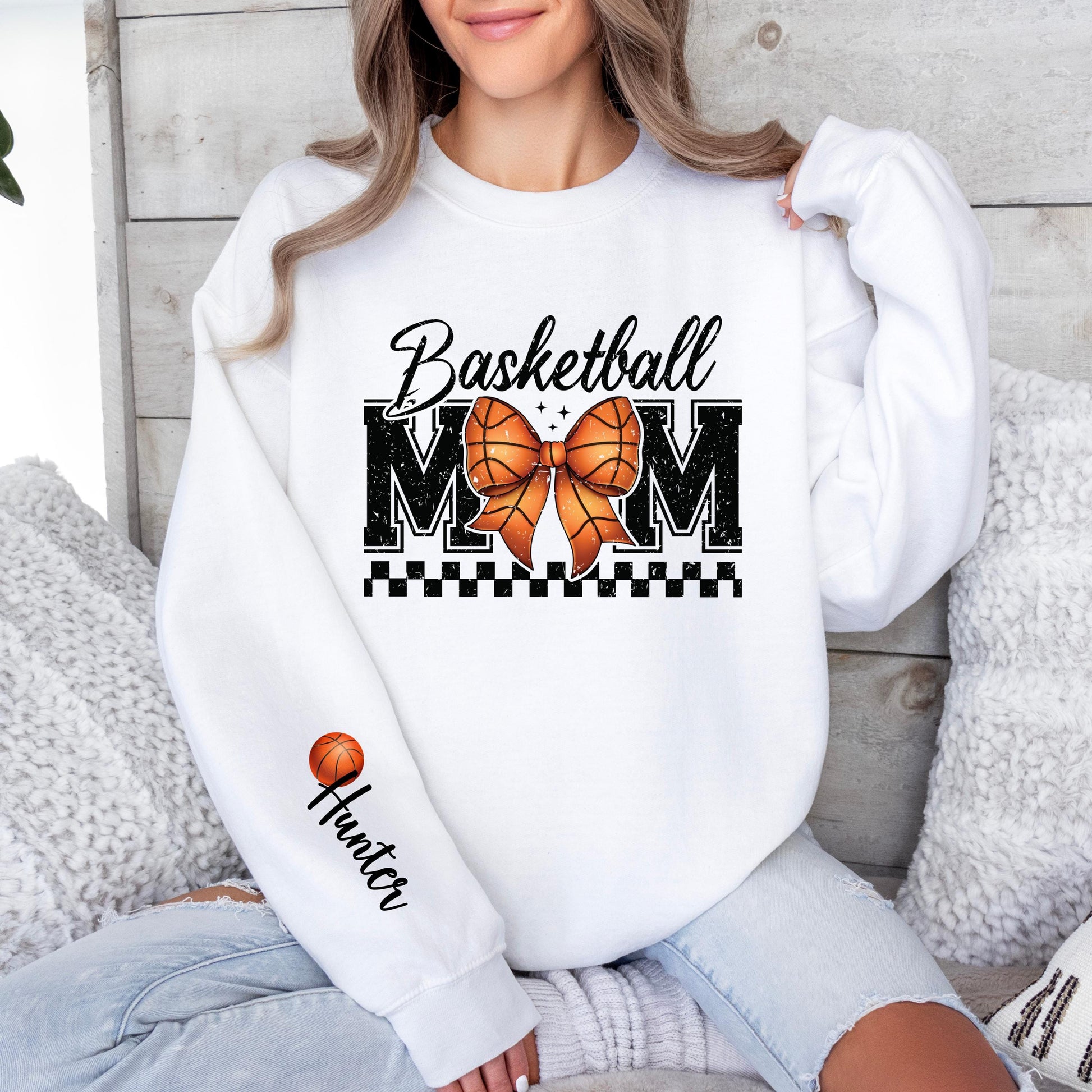 A light gray sweatshirt with "Basketball MOM" in bold black letters, a Basketball-patterned bow in the center, a black checkered underline, and a Basketball icon with "Custom name" written on the sleeve.