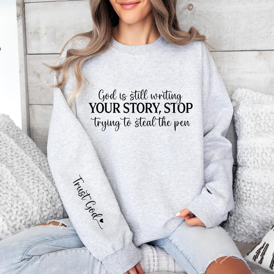 sweatshirt featuring the inspirational quote, &quot;God is still writing your story, stop trying to steal the pen,&quot; in a mix of elegant and bold fonts. The sleeve has &quot;Trust God&quot; written with a small heart.