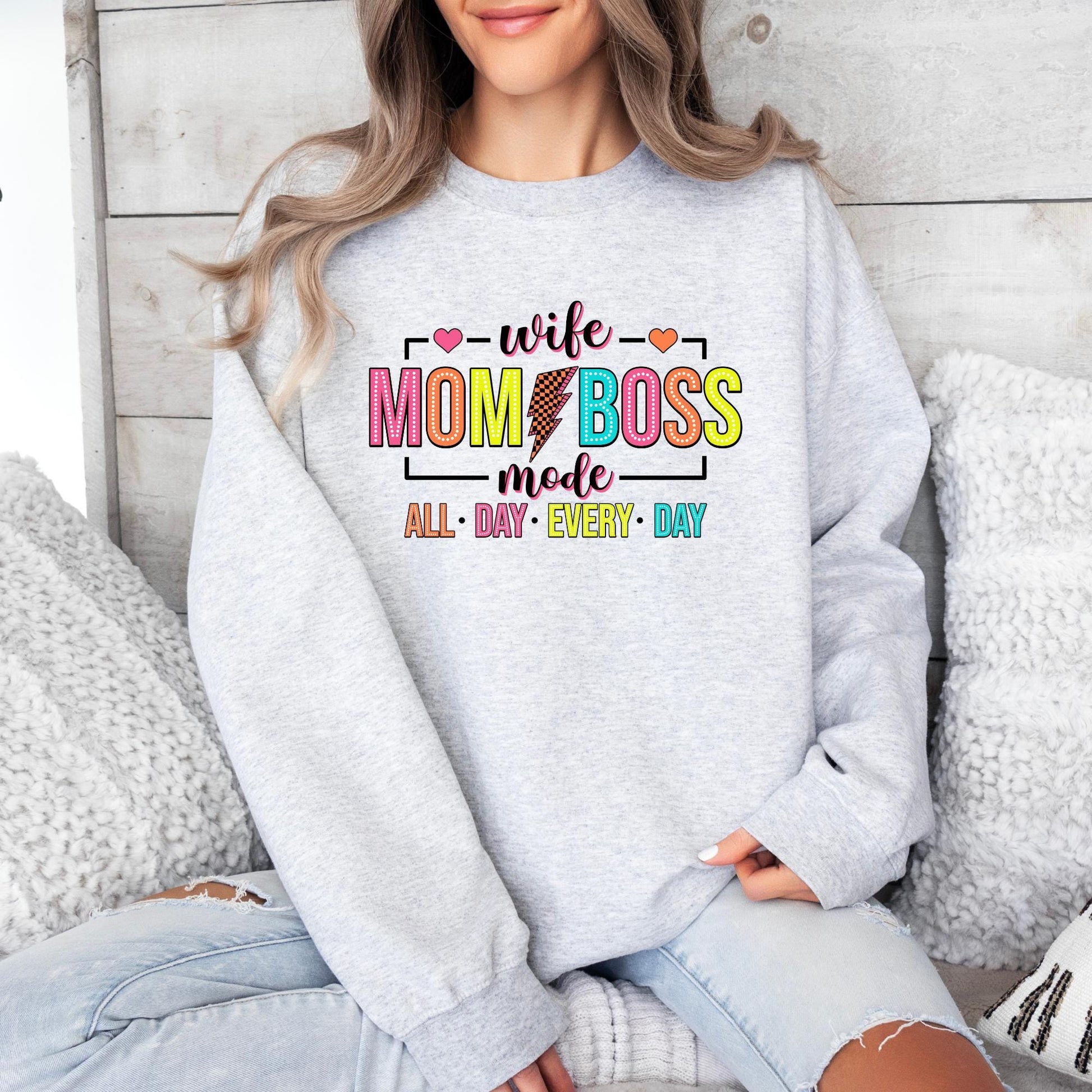 White sweatshirt with &quot;Wife Mom Boss Mode&quot; in bold, colorful letters, featuring a lightning bolt and hearts. The phrase &quot;All Day Every Day&quot; is in bright, playful colors for a fun and empowering look.