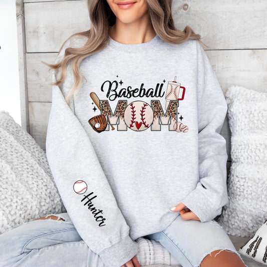 A sweatshirt with a &quot;Baseball MOM&quot; design on the front. The word &quot;MOM&quot; is styled with a baseball pattern, leopard print, a bat, glove, and baseballs. above &quot;MOM,&quot; and there is a baseball-themed drink cup next to the design. The left sleeve features a customizable name area next to a small baseball graphic.