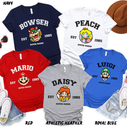 A set of five colorful T-shirts featuring Super Mario characters: Mario (red), Luigi (blue), Peach (white), Daisy (gray), and Bowser (navy). Each shirt has "EST 1985" and "Super Mario" text.
