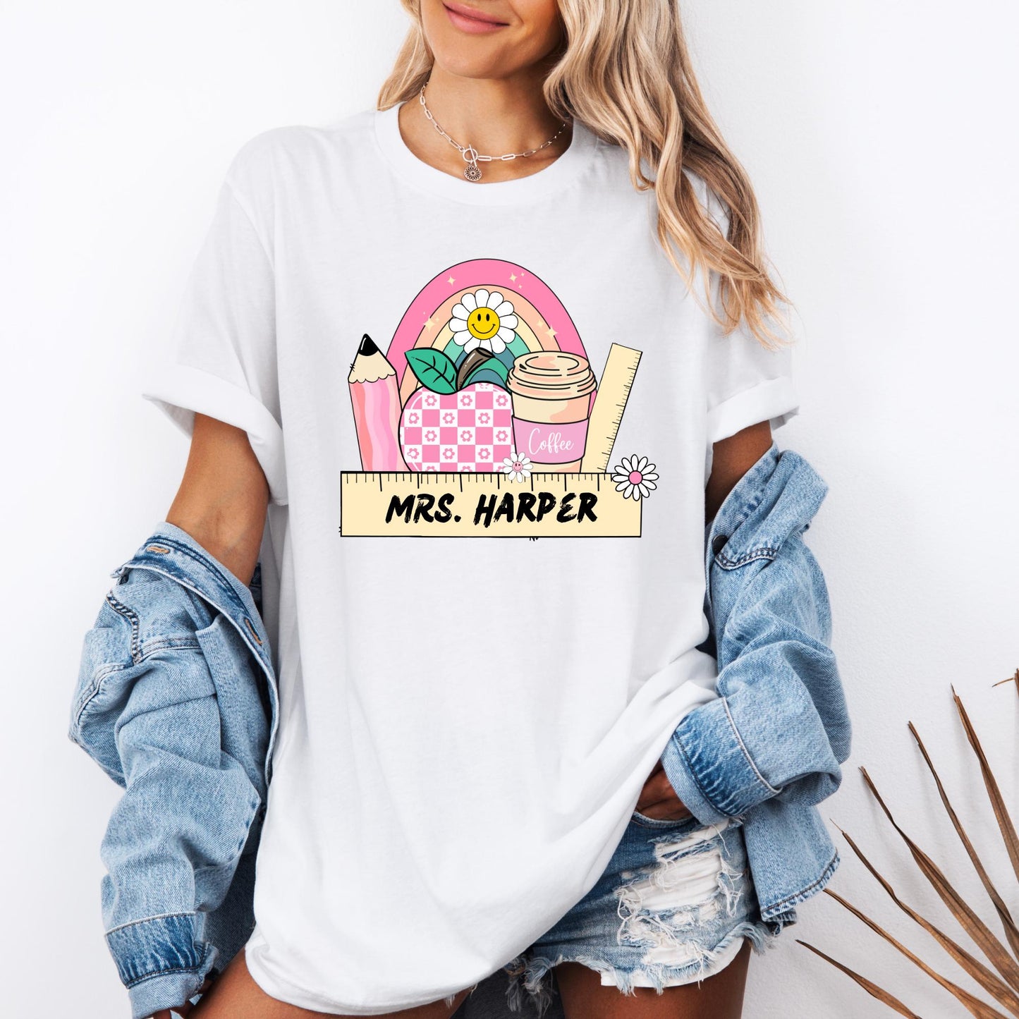 ALT text: A black T-shirt featuring a colorful, teacher-themed graphic design. The design includes a pink pencil, a checkered pink apple, a cup labeled "Coffee," a ruler, green leaves, and a rainbow with a smiling daisy at the center. Below the illustration, a wooden ruler banner displays custom teacher name in bold, black letters, with a small daisy on the right side. The design incorporates pastel tones, creating a fun and cheerful look for educators.
