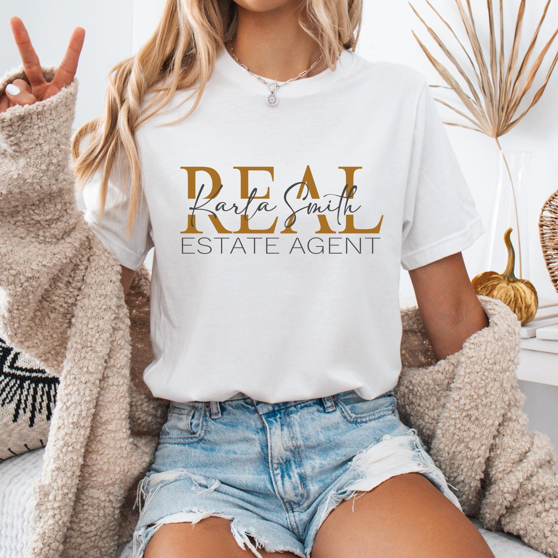 T-shirt featuring a design that reads "REAL ESTATE AGENT" in bold, gold and black uppercase letters. The custom real esta name written in an elegant cursive script overlaying the word "REAL."