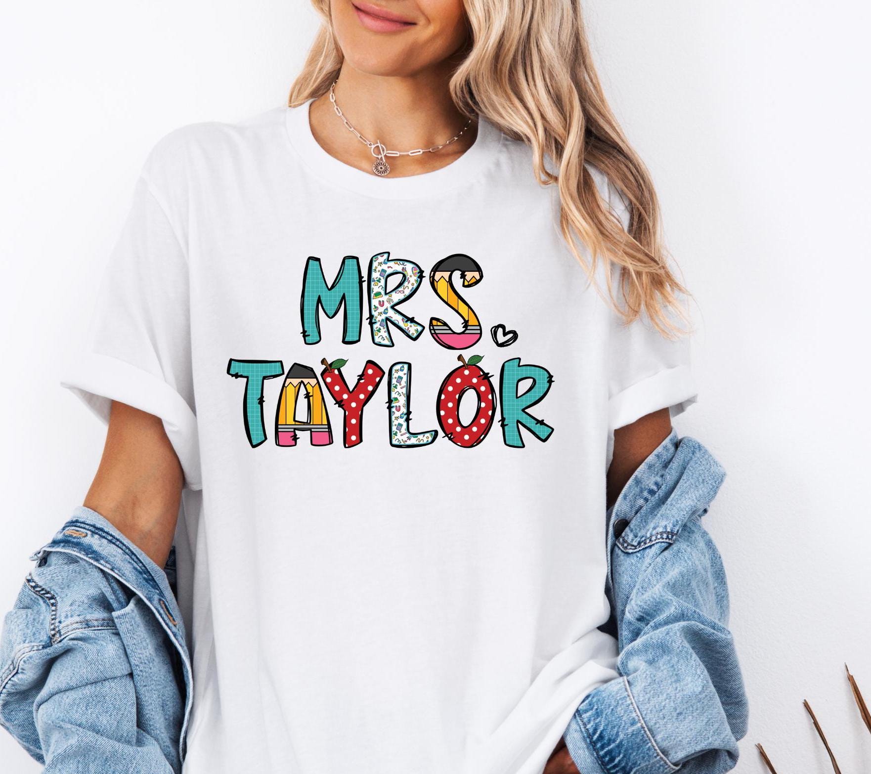 A white T-shirt featuring a colorful, custom teacher name design. The custom teacher name in playful, bold letters, each filled with different school-themed patterns, including polka dots, apples, pencils, and leopard print. A small heart accent is placed next to the text, adding a cute and fun touch. The design is vibrant and perfect for teachers looking for a personalized and stylish outfit.