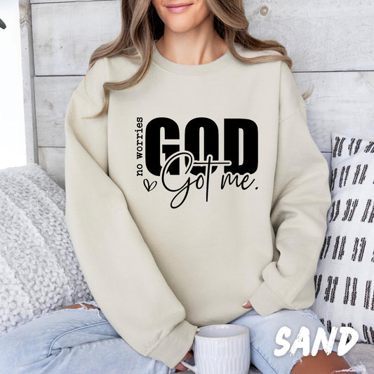 sweatshirt with the phrase &quot;no worries GOD Got me.&quot; printed on the front in bold and script fonts. The design includes a small heart and uses a mix of vertical and horizontal text layout. The woman is sitting comfortably with a mug in hand, dressed casually in ripped light-wash jeans.