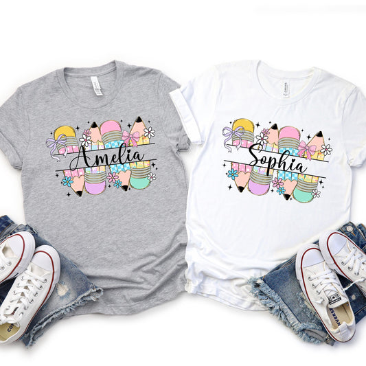 Two girly back-to-school shirts featuring a colorful design of pastel pencils, bows, and flowers, with space to customize the shirt with the child’s name. Shown on gray and white t-shirts, these personalized shirts are perfect for a cute and fun first day of school look.