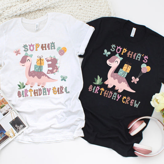 Custom birthday girl t-shirt design featuring a cute pink dinosaur carrying gifts and balloons, surrounded by butterflies and greenery. Includes customizable name and age, with matching "Birthday Girl" and "Birthday Crew" shirt options for family and friends.