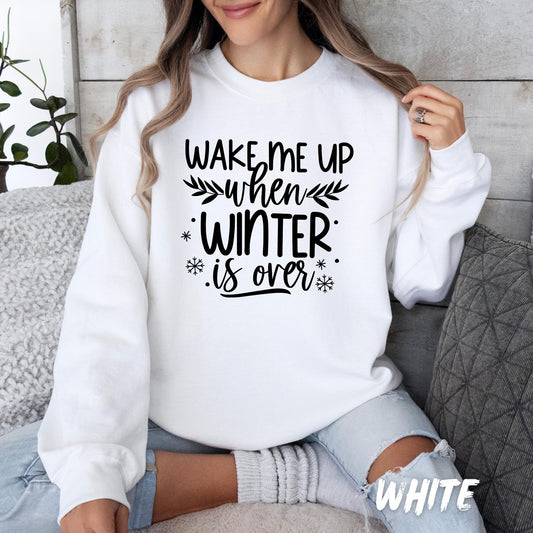 White unisex sweatshirt with black typography design that reads “Wake Me Up When Winter is Over,” featuring snowflakes and leaf accents. A cozy and humorous winter-themed pullover available for adults and kids.