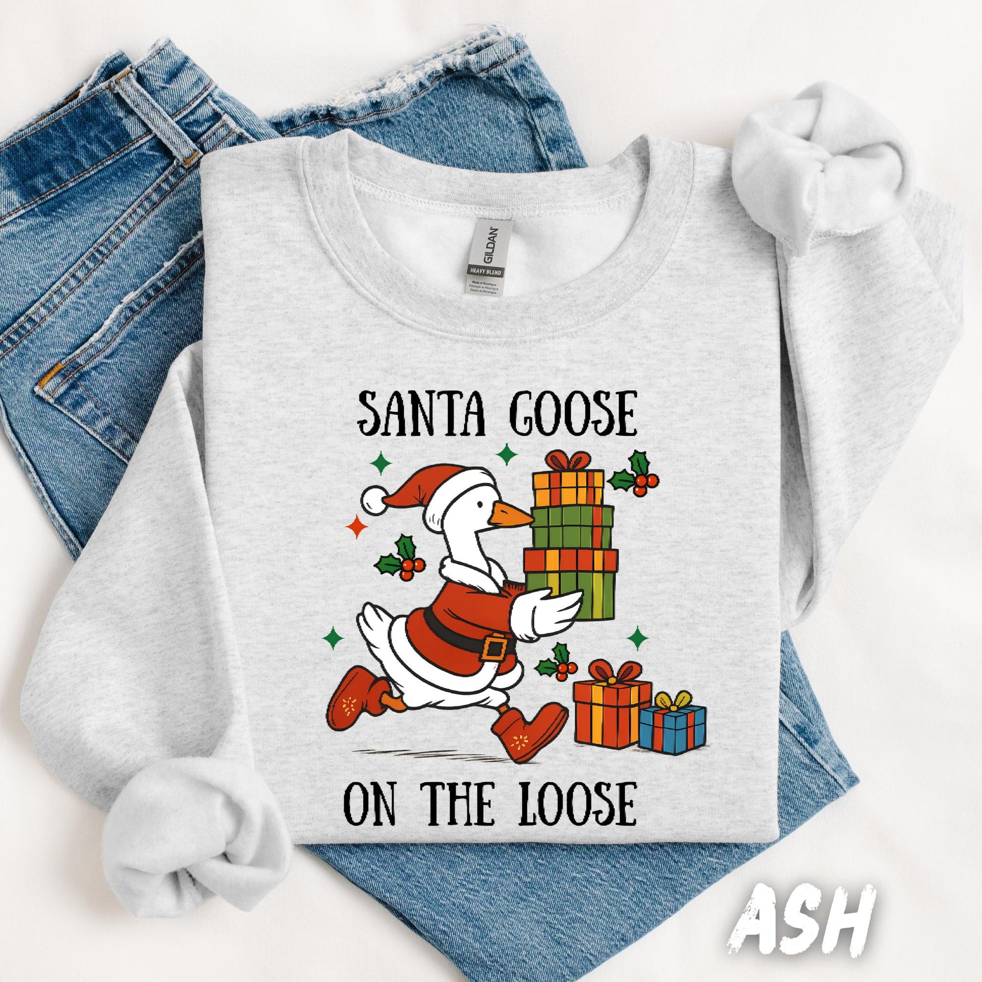 Ash gray unisex sweatshirt featuring a fun design of a goose dressed as Santa running with Christmas presents. Surrounded by holly, stars, and the phrase “Santa Goose On The Loose” in playful black text. A quirky and festive holiday pullover.