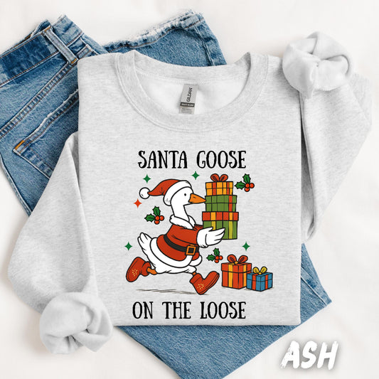 Ash gray unisex sweatshirt featuring a fun design of a goose dressed as Santa running with Christmas presents. Surrounded by holly, stars, and the phrase “Santa Goose On The Loose” in playful black text. A quirky and festive holiday pullover.