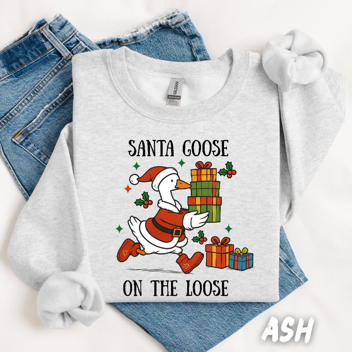 Ash gray unisex sweatshirt featuring a fun design of a goose dressed as Santa running with Christmas presents. Surrounded by holly, stars, and the phrase “Santa Goose On The Loose” in playful black text. A quirky and festive holiday pullover.