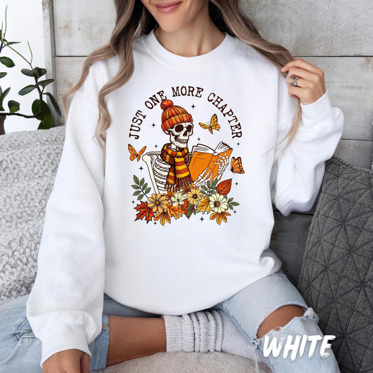sweatshirt featuring a whimsical fall-themed design with a skeleton wearing a striped scarf and knit beanie, holding an orange book. The skeleton is surrounded by autumn flowers, leaves, butterflies, and the phrase &#39;Just One More Chapter&#39; in curved lettering overhead.