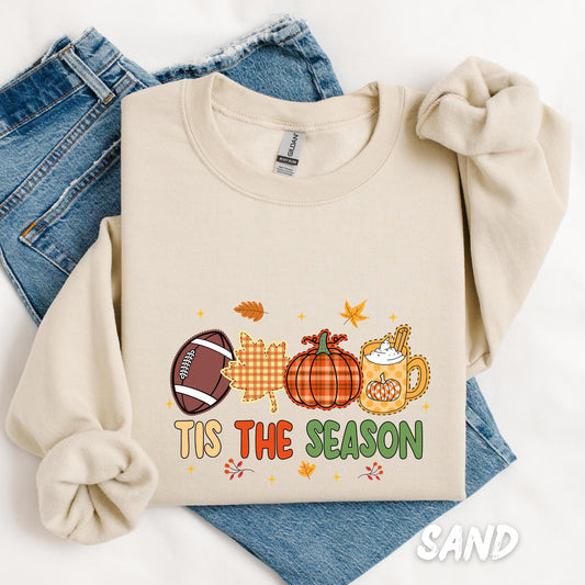 “Tis the Season” sweatshirt featuring a playful fall-themed design with a football, gingham leaf, pumpkin, and a festive mug of pumpkin spice latte, surrounded by autumn leaves and stars.
