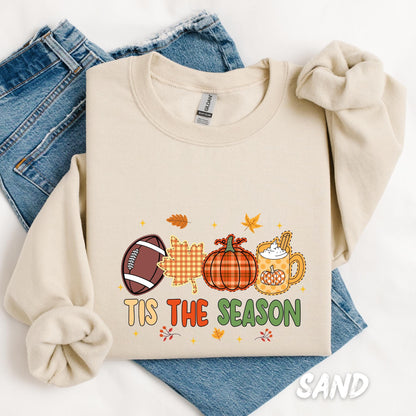 “Tis the Season” sweatshirt featuring a playful fall-themed design with a football, gingham leaf, pumpkin, and a festive mug of pumpkin spice latte, surrounded by autumn leaves and stars.