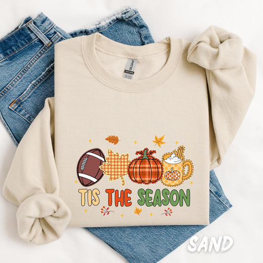 “Tis the Season” sweatshirt featuring a playful fall-themed design with a football, gingham leaf, pumpkin, and a festive mug of pumpkin spice latte, surrounded by autumn leaves and stars.