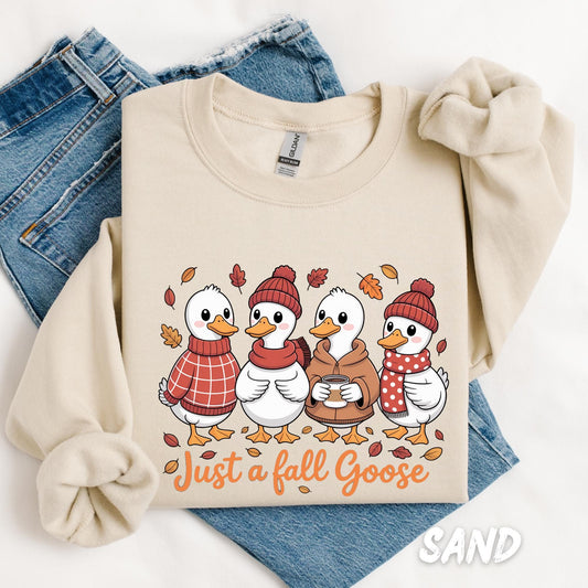 Unisex sweatshirt with a whimsical fall-themed design of four cartoon geese dressed in scarves, sweaters, and beanies. Surrounded by autumn leaves and the phrase “Just a Fall Goose” in orange script, the cozy crewneck is laid over folded jeans.