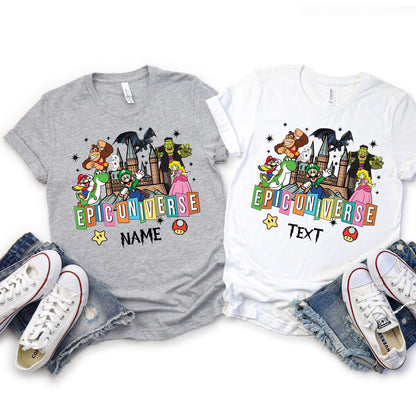 t-shirts, one gray and one white, with a colorful 'Epic Universe' design featuring popular cartoon and video game characters including Mario, Luigi, Yoshi, Princess Peach, Donkey Kong, Frankenstein, and others, standing in front of a castle. The design also includes stars, mushrooms, and customizable text options below the logo.