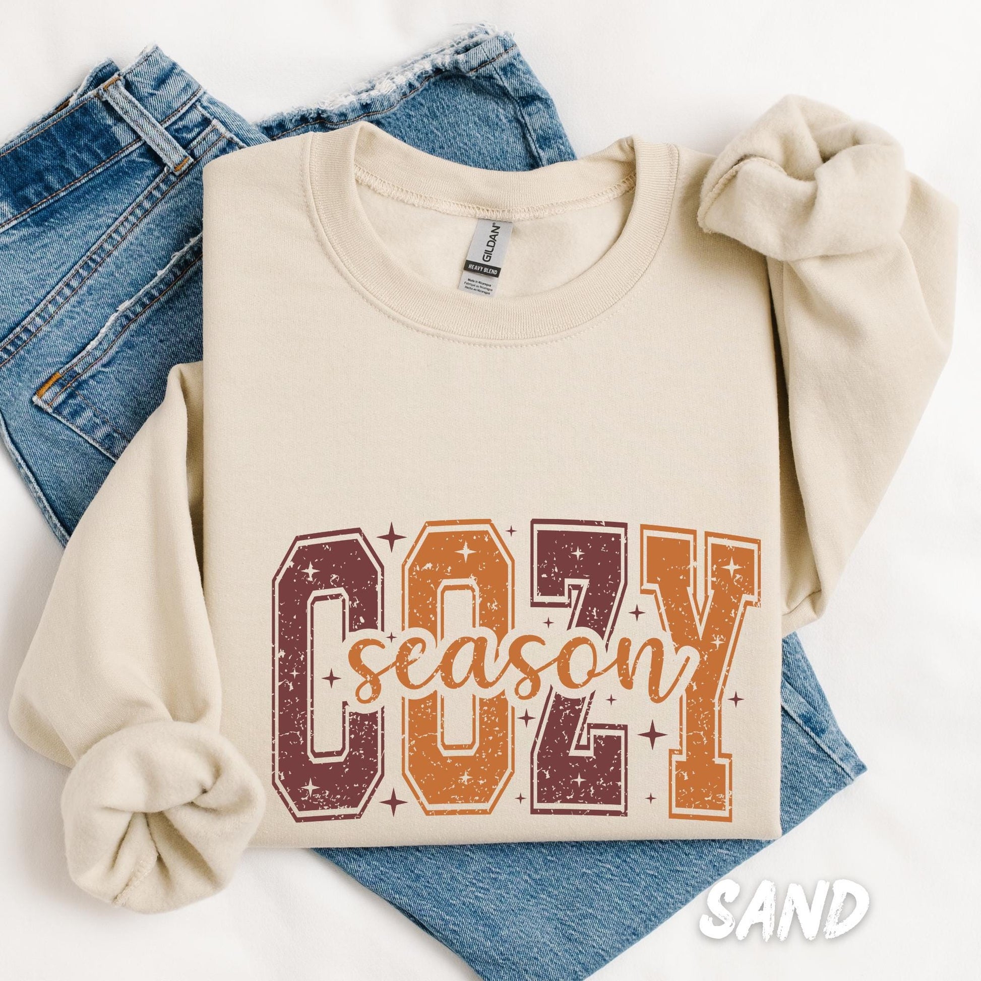 sweatshirt with the phrase "Cozy Season" in large varsity-style letters in autumn colors (burgundy, orange, and golden yellow) with star accents, laid over a pair of folded blue jeans.
