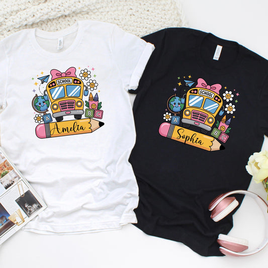 Custom back-to-school t-shirts featuring a colorful school bus design with fun elements like pencils, globes, and ABC blocks. Each shirt includes a large pencil graphic personalized with the child’s name. Available in black and white shirt options. Perfect for kids&#39; first day of school photos and matching classroom outfits.