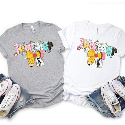 Custom teacher shirt featuring the word &quot;Teacher&quot; in colorful, playful school-themed letters with a yellow pencil bow and customizable name tag – perfect back-to-school or teacher appreciation gift.