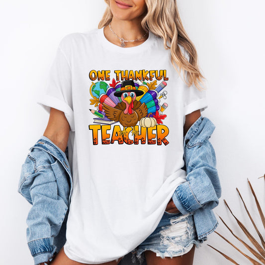 unisex t-shirt featuring a colorful Thanksgiving design with the phrase &quot;One Thankful Teacher.&quot; The graphic shows a cheerful cartoon turkey wearing a pilgrim hat, surrounded by vibrant feathers, autumn leaves, school supplies, and a pumpkin. The word “TEACHER” is printed in bold, festive orange letters, making it a fun and seasonal shirt perfect for educators celebrating Thanksgiving.