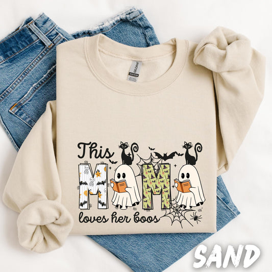 sweatshirt with a Halloween-themed design that reads &quot;This Mom loves her boos.&quot; The word &quot;MOM&quot; is filled with Halloween patterns including ghosts, bats, stars, and witches&#39; hats. Two cute cartoon ghosts, each holding a book, stand over the letters, with black cats perched above and spider webs, bats, and stars completing the spooky, playful look.