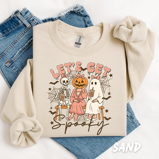 sweatshirt featuring a Halloween-themed design with three cute characters sitting on a bench: a skeleton with a pink bow, a pumpkin-headed figure in a pink dress, and a ghost with a pink bow. Each character holds a drink cup. The text above them reads “LET’S GET” in pink bubble letters, and “spooky” is written in cursive underneath. The design is decorated with spiderwebs, stars, and small bats.