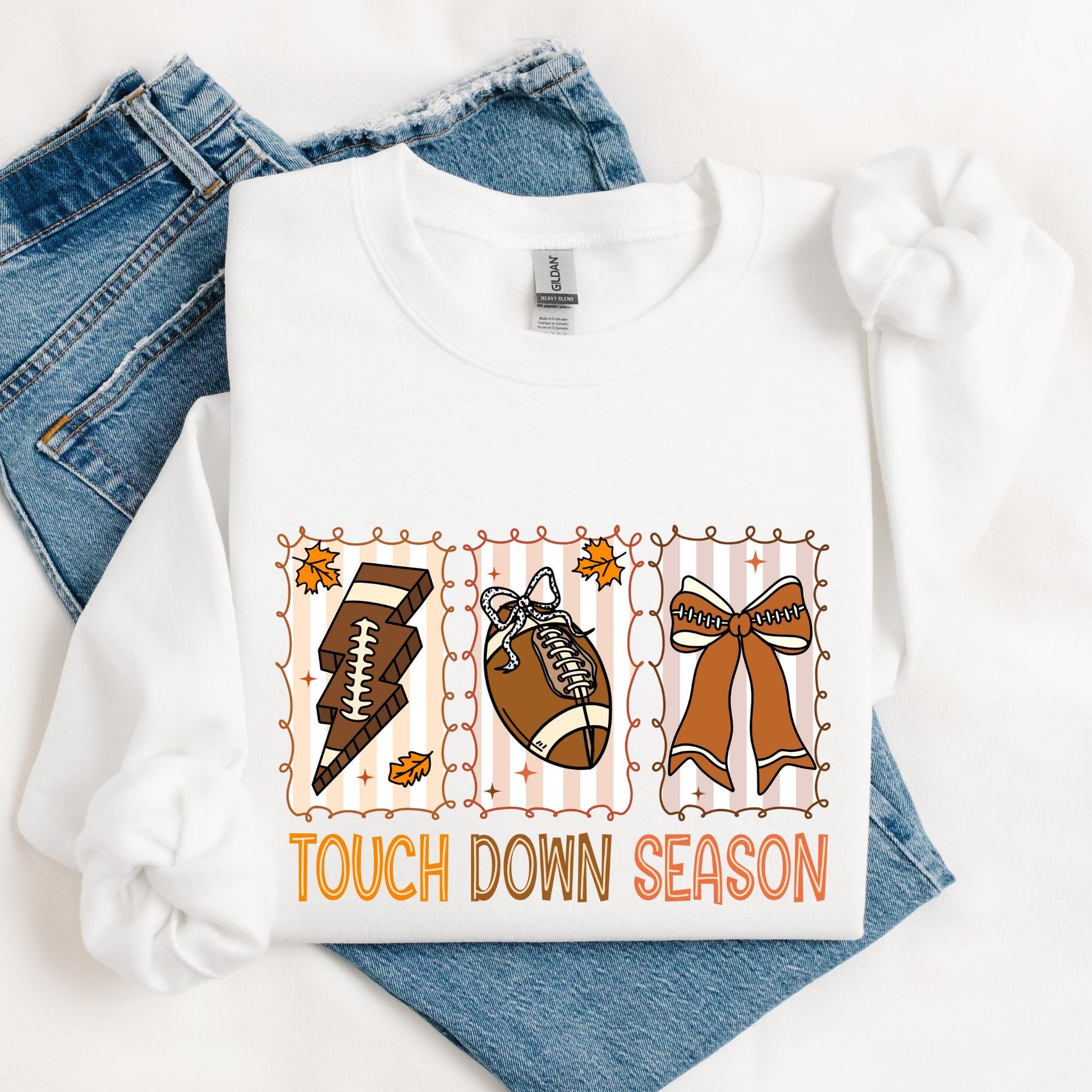 sweatshirt featuring a playful fall-themed football design with three illustrated panels. Each panel includes a football-related item: a lightning bolt made of football laces, a football with a bow, and a large cheer bow. Autumn leaves and swirly frames surround the graphics. Below, the phrase “TOUCH DOWN SEASON” is written in bold orange and brown letters. The shirt is styled with rolled sleeves and laid over a pair of blue denim jeans.