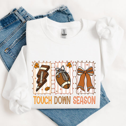 sweatshirt featuring a playful fall-themed football design with three illustrated panels. Each panel includes a football-related item: a lightning bolt made of football laces, a football with a bow, and a large cheer bow. Autumn leaves and swirly frames surround the graphics. Below, the phrase “TOUCH DOWN SEASON” is written in bold orange and brown letters. The shirt is styled with rolled sleeves and laid over a pair of blue denim jeans.