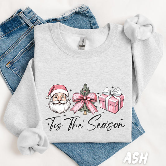 unisex sweatshirt featuring a festive design with Santa Claus, a Christmas tree with a pink bow, and a pink gift box. Text reads “’Tis The Season” in elegant script, surrounded by sparkles. A cute and cozy pullover perfect for holiday vibes.