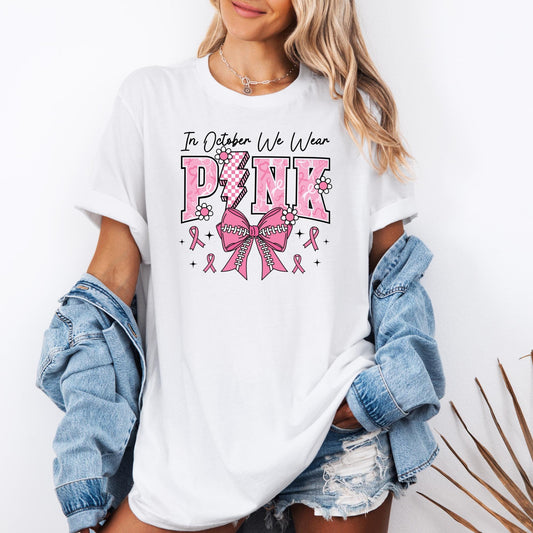 t-shirt with the phrase "In October We Wear Pink" in bold pink letters decorated with flowers, a lightning bolt, and a large pink bow with baseball stitching at the center, surrounded by small pink awareness ribbons and stars. The design supports Breast Cancer Awareness Month.