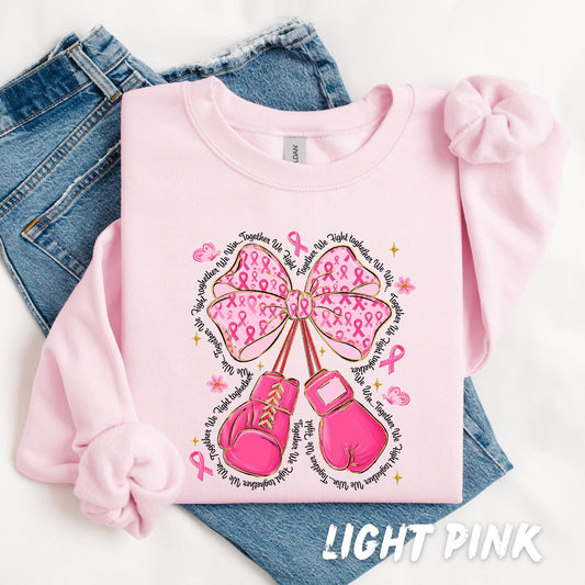 Pink sweatshirt featuring a large bow made of pink awareness ribbons, pink boxing gloves hanging from the bow, surrounded by motivational text that reads “Together We Fight.” The design includes flowers, sparkles, and awareness ribbons. Displayed folded with light denim jeans in the background.