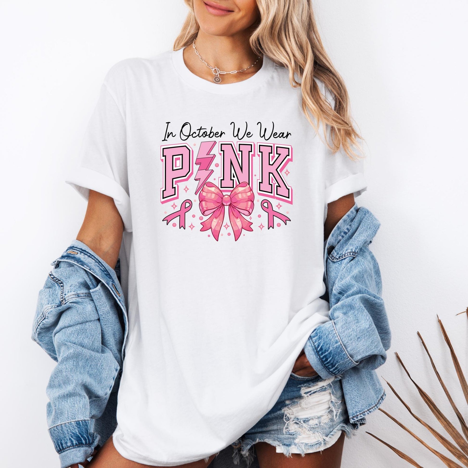 t-shirt featuring the phrase “In October We Wear Pink” printed in bold letters across the chest. The word PINK is highlighted in large pink block letters, with a lightning bolt replacing the “I” and a shiny pink ribbon bow in the center. Small pink breast cancer awareness ribbons and sparkles surround the design, emphasizing the theme of Breast Cancer Awareness Month.