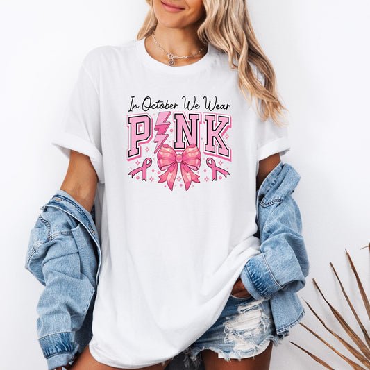 t-shirt featuring the phrase “In October We Wear Pink” printed in bold letters across the chest. The word PINK is highlighted in large pink block letters, with a lightning bolt replacing the “I” and a shiny pink ribbon bow in the center. Small pink breast cancer awareness ribbons and sparkles surround the design, emphasizing the theme of Breast Cancer Awareness Month.