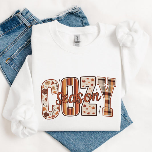 sweatshirt with “Cozy Season” design in large bold letters, each letter filled with autumn-themed patterns like florals, stripes, leaves, and plaid in warm brown, orange, and tan tones.