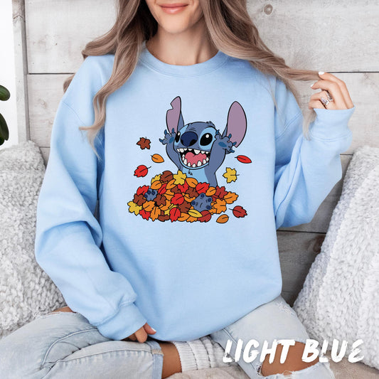 sweatshirt featuring a joyful Stitch character from Disney&#39;s Lilo & Stitch, playfully popping out of a colorful pile of autumn leaves in shades of red, orange, and yellow. The design captures a fun and cozy fall vibe, perfect for seasonal wear.