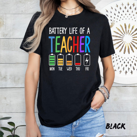 t-shirt featuring the phrase 'Battery Life of a Teacher' in bold white and colorful text. Below, five battery icons represent the week: Monday full green, Tuesday yellow, Wednesday half orange, Thursday low red, and Friday empty with a lightning bolt. The word 'TEACHER' is printed in rainbow colors."