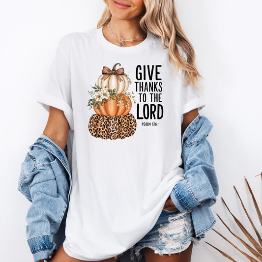 t-shirt with three stacked pumpkins: a leopard-print pumpkin at the bottom, an orange pumpkin in the middle decorated with white flowers, and a cream pumpkin with a brown bow on top. Beside the pumpkins, the text reads “GIVE THANKS TO THE LORD – Psalm 136:1.”