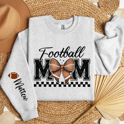 A light gray sweatshirt with &quot;Football MOM&quot; in bold black letters, a football-patterned bow in the center, a black checkered underline, and a football icon with &quot;Custom Name&quot; written on the sleeve.