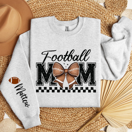 A light gray sweatshirt with &quot;Football MOM&quot; in bold black letters, a football-patterned bow in the center, a black checkered underline, and a football icon with &quot;Custom Name&quot; written on the sleeve.
