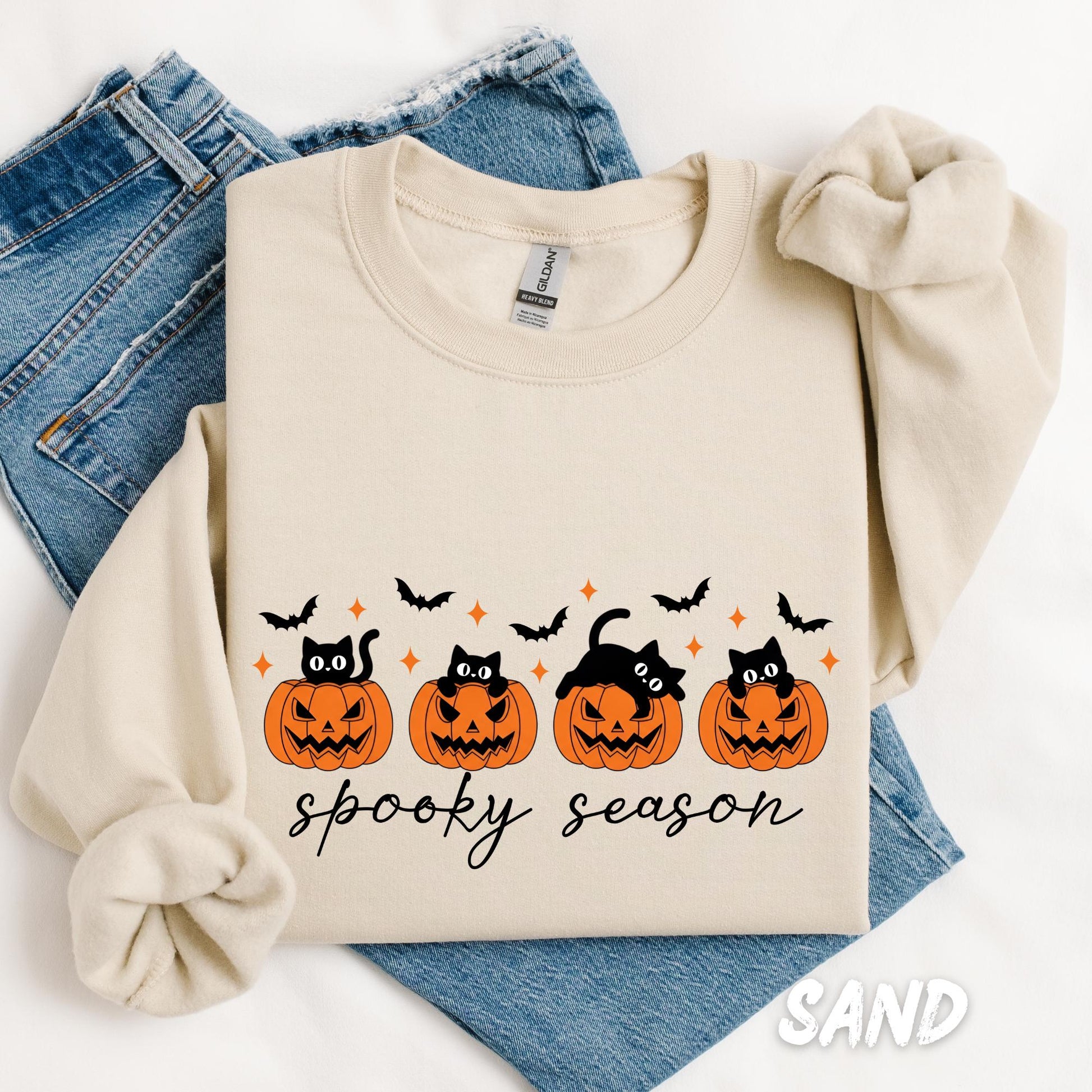sweatshirt folded on a white background with blue jeans underneath. The design features four black cats sitting inside or peeking out of orange jack-o'-lantern pumpkins, surrounded by flying black bats and small sparkles. Below the pumpkins, the text "spooky season" is written in a stylish cursive font.