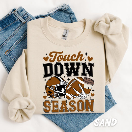 sweatshirt with a bold &quot;Touch Down Season&quot; design in brown and black. The graphic features a football helmet, football, skeleton hand, and coffee cup with heart and star accents, capturing the fall football season vibes. The sweatshirt is paired with light-wash denim jeans and laid out flat on a white background.
