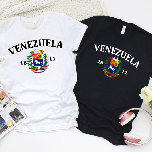 Custom T-shirt design featuring the word “VENEZUELA” in bold black capital letters above the country’s national coat of arms. The coat of arms includes a red, yellow, and blue shield with a white horse, wheat, and weapons, framed by a palm and olive branch. Above the shield is a ribbon in the Venezuelan flag colors (yellow, blue, and red). Below the design, the numbers “18” and “11” are displayed on either side, representing the year 1811.